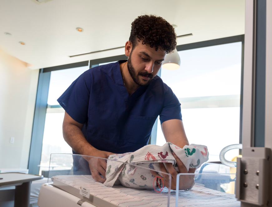 medical student in scrubs placing an infant wrapped in a blanket down on a bed