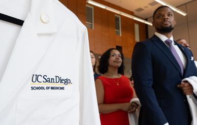 close up photo of white coat with school of medicine logo on it. Black male student in background