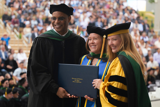 student in commencement regalia  flanked by male dean on left and female dean on right